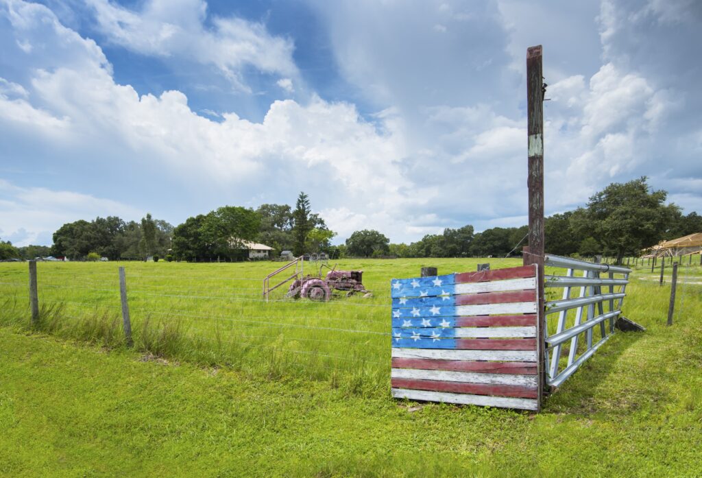 Beneath a blue sky with big white fluffy clouds, a corner of a fence in a green field is painted to look like the American flag, with red and white stripes and white stars on a blue background.